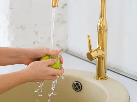Hands washing a green apple under a gold faucet