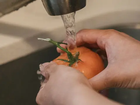 Hands rinsing a tomato under a kitchen faucet