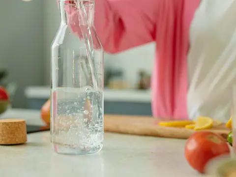 A glass bottle being filled with water on a kitchen counter beside lemon slices