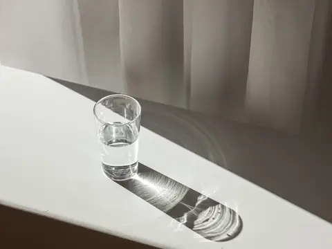 A glass of water on a white table casting shadows in soft sunlight