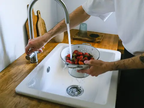 Water running from a kitchen faucet over strawberries in a colander in a white sink